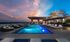 Rooftop pool overlooking the city at night with a cabana, white, brown, and blue pool lounge chairs, and trees surrounding the area.