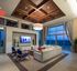 Lounge with floor-to-ceiling windows, brown paneled ceiling with a beige chandelier, white stone wall with a flat-screen TV, and a white sectional couch with blue chairs.
