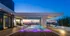 An outdoor lap pool deck with a wide overhang shading part of the pool. There are purple underwater lights shining on 4 in-pool lounge chairs, marble-clad landscaping boxes with large bushes, and a sunset view of the downtown Houston skyline.