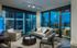 Spacious living room with two walls of full-width floor-to-ceiling windows allowing for a panoramic city view. The room has a couch, coffee table, three armchairs, media console, dark wood-style floors, and a door to a balcony.