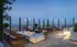 Multiple seating areas placed at the corner of the rooftop with glass walls allowing for a panoramic view of the city. There are 6 armchairs around a minimalist marble fire pit, a patio table and chairs, and landscaping throughout.