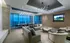 Lounge area with a 12-person dining table, a seating area with two couches, and two armchairs, a large HDTV mounted on the wall, high ceilings, and a full-width floor-to-ceiling window with a city skyline view.