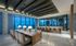 Resident lounge with an 8-person table for dining or work, a work area and wet bar with 7 chairs along a marble desk, and two full-width floor-to-ceiling windows with a city view. The floors, walls, and columns are marble-clad.