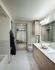 A bathroom with a large tub, marble tiling on the floors and walls, a walk-in closet with beige carpet, and a dual vanity with white quartz counters and handleless light wood cabinets.