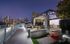 A landscaped rooftop entertaining area with a dining table, grilling station, and several lounge chairs facing a city skyline view.