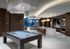 A clubhouse with dark wood-paneled walls, a billiards table under a rectangular chandelier with hanging silver chains, and a resident catering kitchen with two ovens, a large marble island with barstools, and an HDTV.