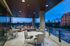 An open-air loggia with a view of the city skyline at dusk, a wraparound glass railing, a seating area with a curved couch and armchairs, and multiple sets of white tables and chairs.
