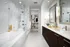 Spacious bathroom with a walk-in glass shower, large soaking tub, white marble walls and flooring, dark wood cabinetry and a large two-sink vanity with a white quartz countertop. There is an entrance to a walk-in closet.