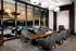 Lounge with a dark wood table and grey upholstered chairs, floor-to-ceiling windows facing the pool deck. Decorative plants and artwork line the room.