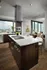 Open-concept kitchen with high ceilings, stainless steel appliances, modern dark wood cabinetry, a white quartz island with three barstools and light wood style floors. There is an exterior window above the sink, and the room opens to a living room with floor-to-ceiling windows.