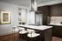 Kitchen with wood-style floors, dark wood cabinetry, stainless steel appliances, white quartz countertops, and an island with three white barstools. There is artwork on the wall next to an entrance to a laundry room.