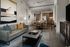 Open living room with grey, blue and dark wood furnishings. The living room opens to a dining area with a light wood table and chairs, next to a built-in desk. The dining area opens to a kitchen with light wood cabinetry and stainless steel appliances.