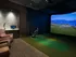 Indoor virtual golf simulator with a green artificial-grass area in front of a large projector screen showing a golf course simulation.