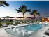 Outdoor pool deck with palm trees, a cabana, lounge chairs on the edge of the pool and in the shallow ends, and a city skyline view.