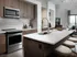 A kitchen with stainless steel appliances, wood cabinetry, a white quartz island with a large sink and three white barstool. There is a dry bar next to the kitchen and an entrance to a living room.