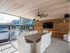 An open-air dining room by the pool enclosed with a gate, with a wood-paneled wall and ceiling, a flat-screen TV mounted on the wall, ten white upholstered chairs, and a ceiling fan.