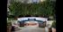 A round banquette couch and two wicker armchairs around a round stone fire pit with trees in the background.