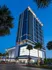 Exterior rendering of The Driscoll at River Oaks high-rise apartment building, with floor-to-ceiling window walls, balconies on some units, a lit up parking garage near the entrance, and palm trees lining the sidewalk.