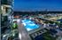 Outdoor pool deck with lounge chairs surrounding the pool, a grass area with white lounge chairs, apartment balconies overlooking the pool, and a city skyline view.