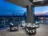 Open outdoor deck with white and grey upholstered chairs seated around a grey coffee table. There is a grey marble pillar and glass walls overlooking the city.