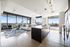 angled view of apartment living and kitchen with floor to ceiling windows showing views of downtown Houston skyline, light wood style flooring, kitchen island with brown cabinets and beige stone counters with 3 pendant lights above