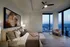 Luxurious bedroom with floor-to-ceiling windows, offering breathtaking city views. Elegant decor featuring a plush bed, modern art, and cozy seating area.