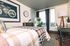 Modern bedroom with a checkered quilt, wall art, and a writing desk, featuring a city view through large windows.