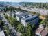 Aerial view of a modern apartment complex nestled in a charming green neighborhood, with a scenic bridge and vibrant city skyline in the background.