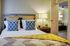 Cozy bedroom with stylish decor, featuring geometric blue pillows, warm lighting, and a view into a modern living area.