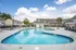 Inviting outdoor swimming pool area with lounge chairs, surrounded by modern apartment buildings and lush greenery under a bright blue sky.