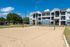 Modern apartment complex with a charming facade, surrounded by lush greenery and featuring a well-maintained sand volleyball court under a vibrant blue sky.
