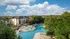 Luxurious apartment complex featuring a large resort-style swimming pool surrounded by lush greenery and elegant seating areas under a bright blue sky.