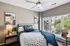 Bright and cozy bedroom with modern decor, featuring a plush bed with blue accents, an elegant ceiling fan, and large windows offering scenic views for ample natural light.