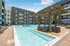 Modern apartment complex featuring a luxurious outdoor pool with stylish seating, surrounded by contemporary buildings and blue umbrellas under a clear sky.