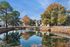 Scenic view of modern apartments by a serene lake surrounded by autumn trees and a white picket fence.