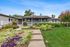 Charming gray bungalow with a welcoming porch, surrounded by vibrant gardens and lush green lawns under a sunny blue sky.
