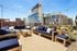 Chic urban rooftop patio with modern blue cushioned seating, wooden tables, and lush greenery, offering stunning cityscape views.