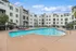 Elegant apartment complex with a sparkling outdoor pool, modern lounges, and lush landscaping under a bright blue sky.