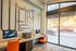 Modern apartment lounge with sleek computer workstations, vibrant orange chairs, and large glass windows offering urban street views.