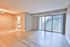 Bright and spacious living room with sleek wooden flooring, large windows with vertical blinds, and a modern kitchen entrance.