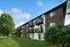 Charming brick apartment building with private balconies, surrounded by lush greenery and a serene lawn, under a clear blue sky.