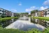 Charming apartment complex with scenic pond view, lush greenery, and elegant architecture under a clear blue sky.