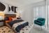 Modern bedroom with elegant decor, featuring a cozy bed with vibrant pillows, stylish abstract artwork, and a chic teal armchair by a sunlit window.