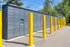Modern outdoor Amazon locker station with a sleek gray design, set against a vibrant backdrop of yellow bollards and lush greenery.