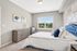 Bright and inviting bedroom with elegant decor, featuring a plush blue headboard, cozy chair by the window, and serene artwork.