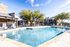 Inviting apartment complex pool area with crystal-clear water, lush greenery, and ample seating under blue umbrellas for a relaxing atmosphere.