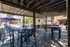 Chic outdoor patio area with modern seating under a wooden pergola, overlooking a sunny, inviting pool with blue umbrellas and surrounded by lush greenery.