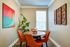 Charming dining nook with vibrant orange chairs, modern wall art, lush green plant, and natural light streaming through blinds, creating a cozy and inviting atmosphere.