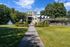Charming landscape view showcasing a scenic pathway leading to a modern clubhouse with a serene fountain and lush greenery in the foreground, framed by tall trees and a towering apartment building under a clear blue sky.