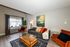 Chic living room with modern decor, featuring a cozy sofa with vibrant throw pillows, elegant glass coffee table, and plush orange chairs, enhanced by natural light from large windows and stylish artwork.